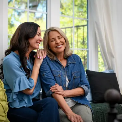 Two women, appearing to be mother and daughter, sitting together on a couch smiling warmly while embracing in a bright living room with large windows