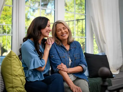 Two women, appearing to be mother and daughter, sitting together on a couch smiling warmly while embracing in a bright living room with large windows