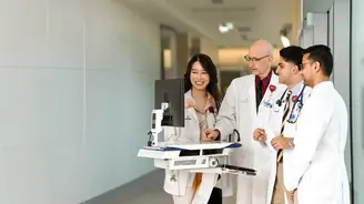 Four healthcare professionals in white coats gather around a mobile computer workstation in a hospital hallway, smiling and talking.