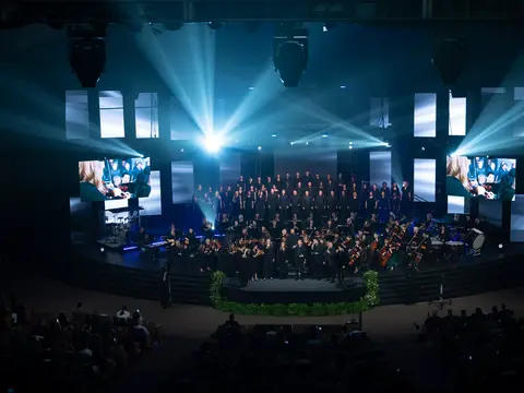 The AdventHealth Choir and orchestra perform on a large blue-lit stage beneath bright spotlights and video screens.