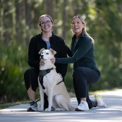 Two women and a dog sit on a sidewalk, smiling at the camera.