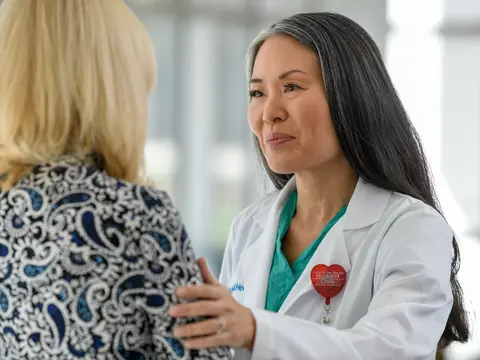 A doctor in a white coat and green shirt talks to a patient in a blue and white patterned shirt.