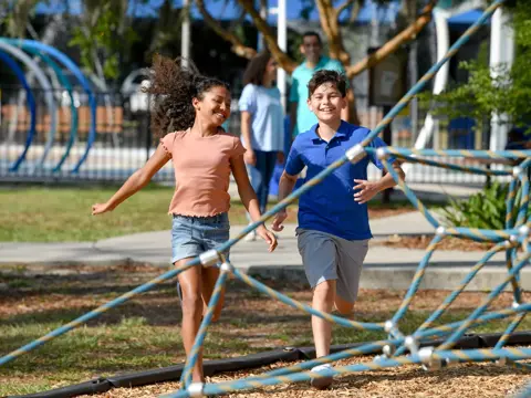 Two children run through a playground, smiling and enjoying the outdoor environment.