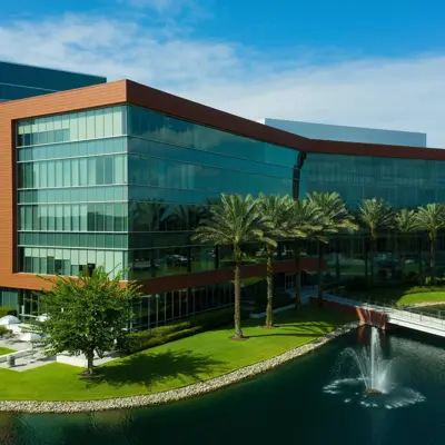 Modern office building with glass facade, surrounded by palm trees and a pond with fountains.