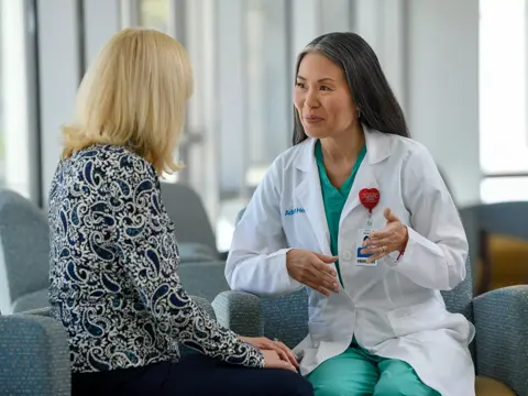 A doctor in a white coat and green scrubs talks to a patient in a waiting room.