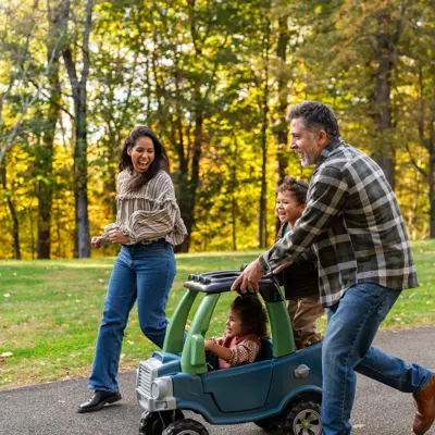 Parents and two children playing together outdoors with a toy car on a paved path in a park with autumn trees in the background