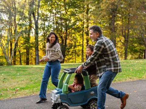 Parents and two children playing together outdoors with a toy car on a paved path in a park with autumn trees in the background