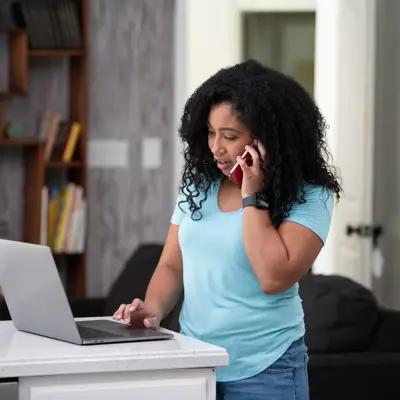 A woman in a blue shirt talks on her phone while working on a laptop at home.