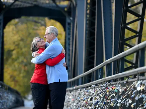 An elderly couple hugging on a bridge with padlocks, symbolizing love and commitment.