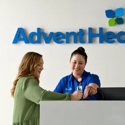 A smiling woman in a blue shirt assists a customer at the AdventHealth reception desk.