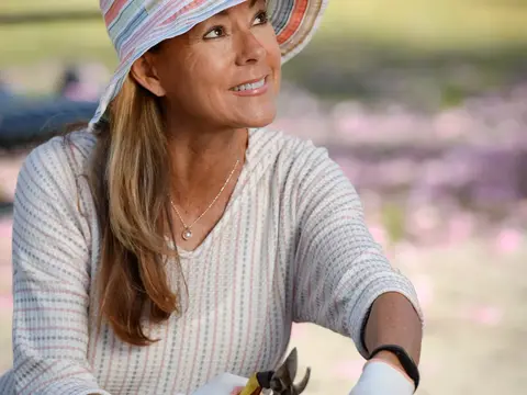 A woman wearing a hat and gloves is trimming purple flowers with pruning shears.