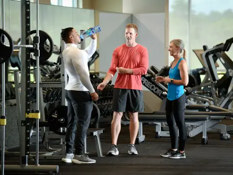 Three people in a gym, one drinking water, engaging in conversation.