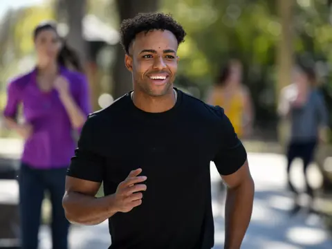 A smiling man jogs down a path with other runners in the background.