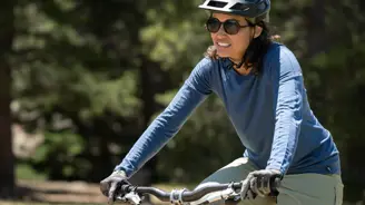A woman wearing a blue shirt and helmet rides a bike outdoors with trees in the background.
