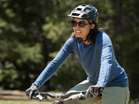A woman wearing a blue shirt and helmet rides a bike outdoors with trees in the background.
