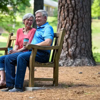 Smiling older couple sitting together on a wooden park bench in an outdoor setting with trees and a gazebo in the background