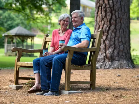 Smiling older couple sitting together on a wooden park bench in an outdoor setting with trees and a gazebo in the background