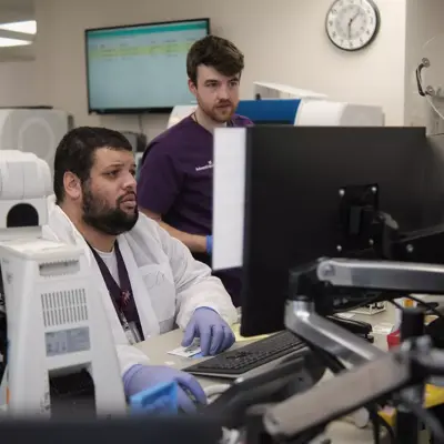 Two men in a lab, one in a white coat working on a microscope, the other in purple observing.