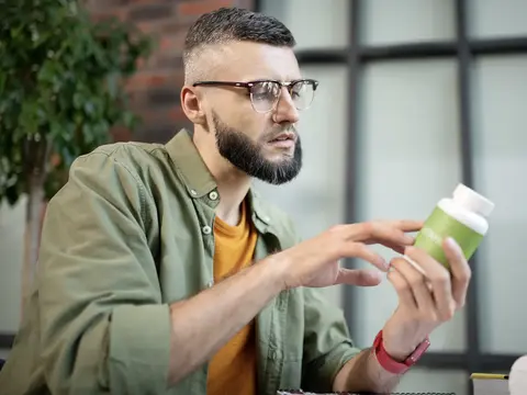 Man with glasses and beard wearing olive green shirt over yellow t-shirt reading the label of a green supplement bottle in a modern office or home setting