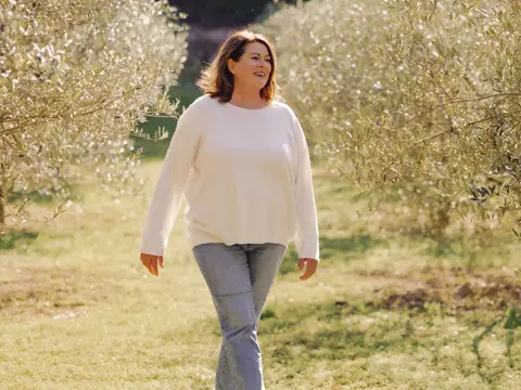 Woman in white sweater walks along a sunlit path between olive trees in an orchard, smiling with a serene expression