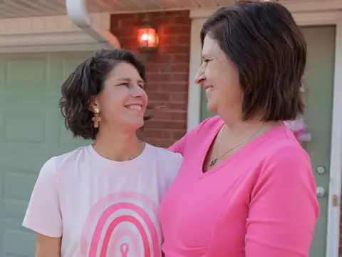 Two women smiling at each other outdoors in front of a brick house with a green door, one wearing a white t-shirt with pink rainbow design and the other in a bright pink top