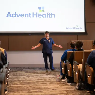 A woman in a blue uniform stands on stage in front of an AdventHealth logo, addressing an audience.