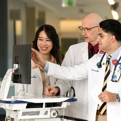 Four medical professionals in white coats gathered around medical equipment, with a senior physician pointing at a monitor while three residents observe and smile in a hospital hallway.
