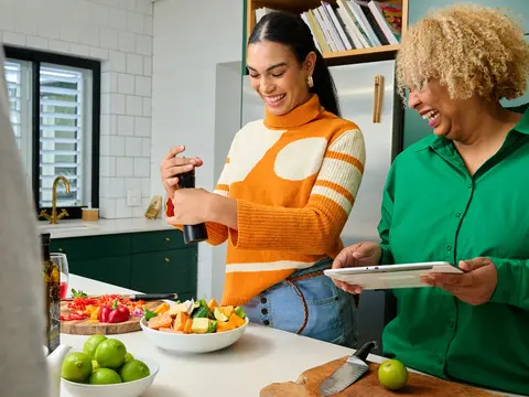 Two people prepare food in a bright kitchen; one pours dressing into a jar while the other holds a plate; vegetables on counter.