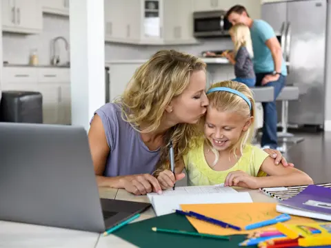 Mother and daughter working on homework at the kitchen table with father in the background.