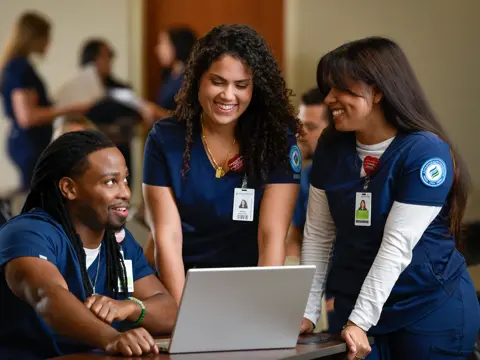 Three smiling students in blue scrubs with AdventHealth University badges gathered around a laptop in a classroom setting, engaged in collaborative learning.