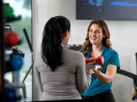 A fitness instructor in a blue shirt holds a red dumbbell while talking to a client in a gym.