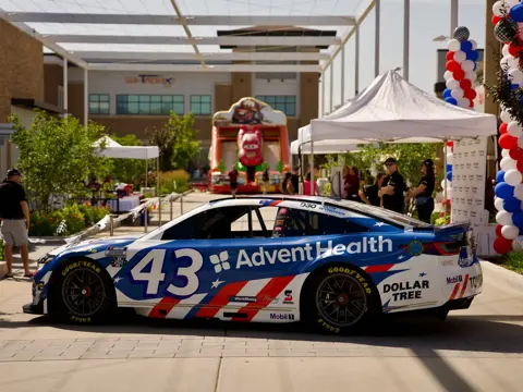 Blue and white race car with AdventHealth branding is parked at an outdoor event with tents, balloon columns, and a racing-themed inflatable in the background.
