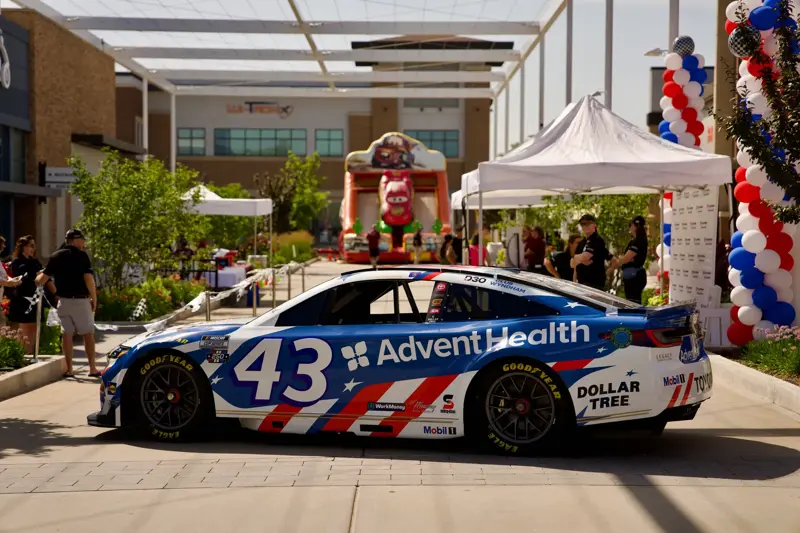 Blue and white race car with AdventHealth branding is parked at an outdoor event with tents, balloon columns, and a racing-themed inflatable in the background.