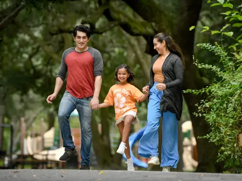 A family of three walks together in a park, smiling and holding hands.