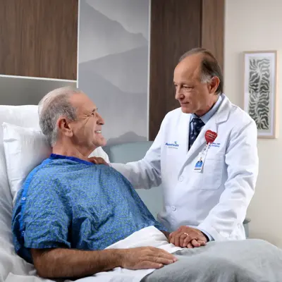 A smiling doctor in a white coat holds the hand of a smiling patient in a hospital bed.