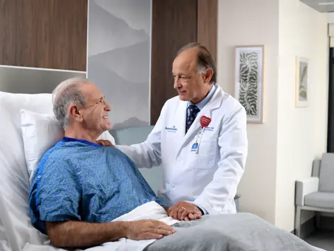 A smiling doctor in a white coat holds the hand of a smiling patient in a hospital bed.