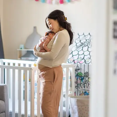 Mother in beige sweater tenderly holding and kissing her newborn baby in a softly lit nursery with a white crib.
