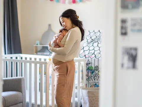 Mother in beige sweater tenderly holding and kissing her newborn baby in a softly lit nursery with a white crib.