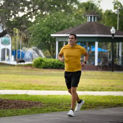 A man in a yellow shirt and black shorts jogs on a sidewalk in a park.
