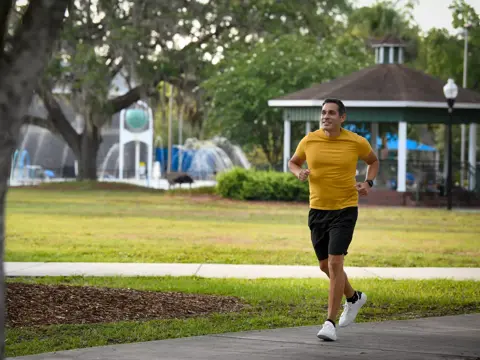 A man in a yellow shirt and black shorts jogs on a sidewalk in a park.