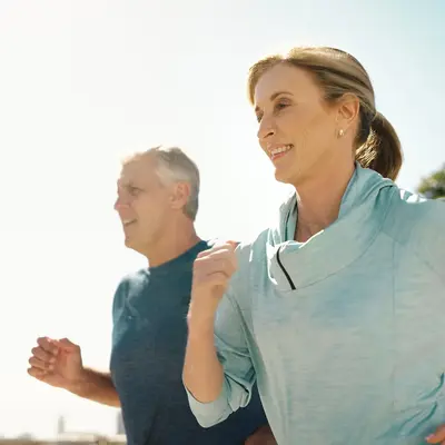 Older couple jogging outdoors in athletic wear, smiling as they run together on a sunny day with trees in the background
