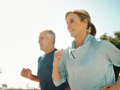Older couple jogging outdoors in athletic wear, smiling as they run together on a sunny day with trees in the background