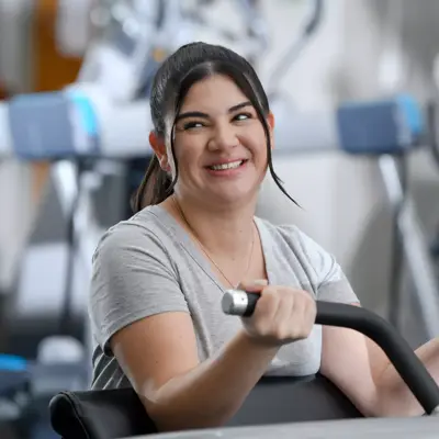 A smiling woman in a gray t-shirt using a weight machine in a gym.