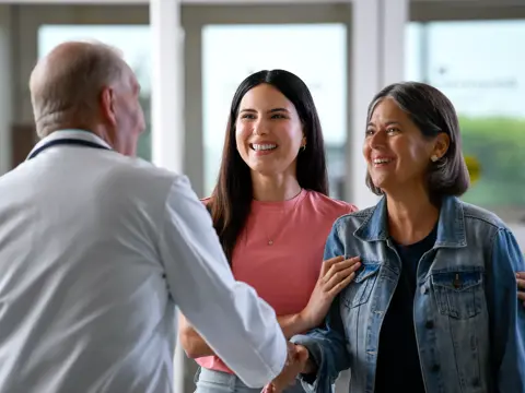 A doctor shakes hands with two smiling women in a hospital corridor.
