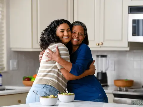 A young woman and an older woman embrace warmly in a bright modern kitchen, smiling at the camera with bowls of salad on the counter in front of them.