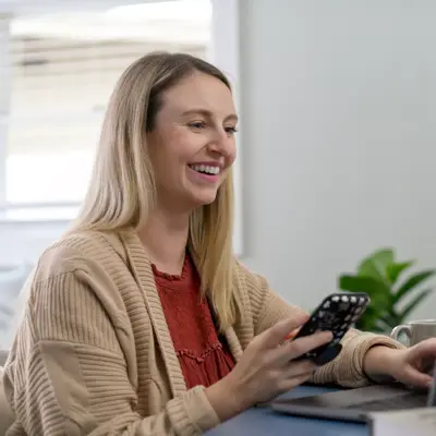 A smiling woman using a smartphone while sitting at a desk with a laptop and candle.