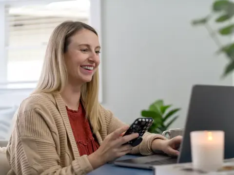 A smiling woman using a smartphone while sitting at a desk with a laptop and candle.