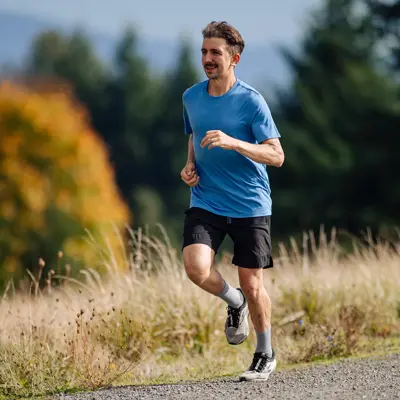 Man jogging on a gravel path through grassy field with trees and mountains in background.