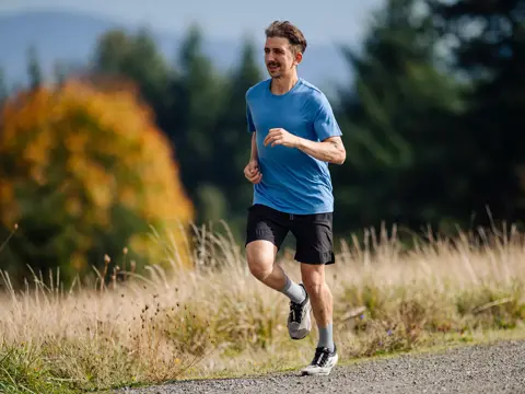 Man jogging on a gravel path through grassy field with trees and mountains in background.