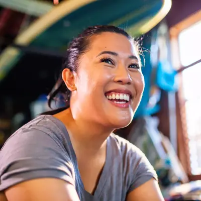 A smiling woman in a gray shirt stands in a room with surfboards and a bright window.